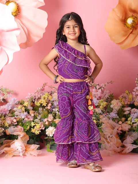 Young girl in a purple traditional outfit standing in front of floral decorations on a pink background