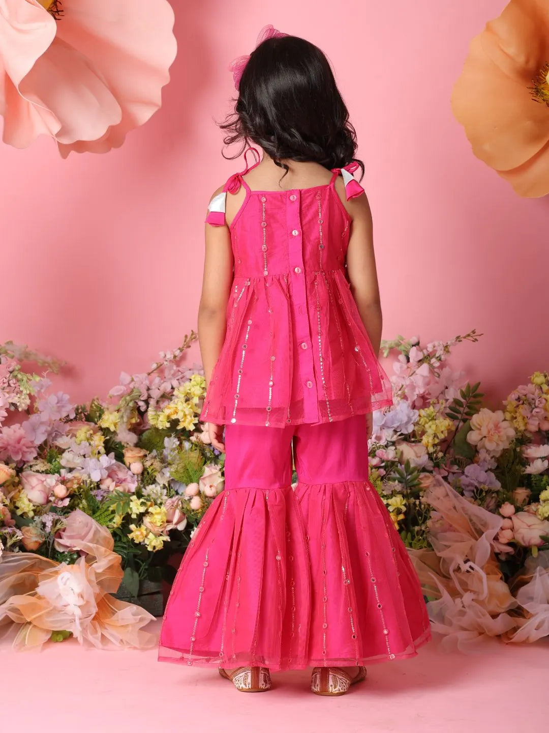 Young girl in a pink traditional outfit standing against a pink background with flowers.