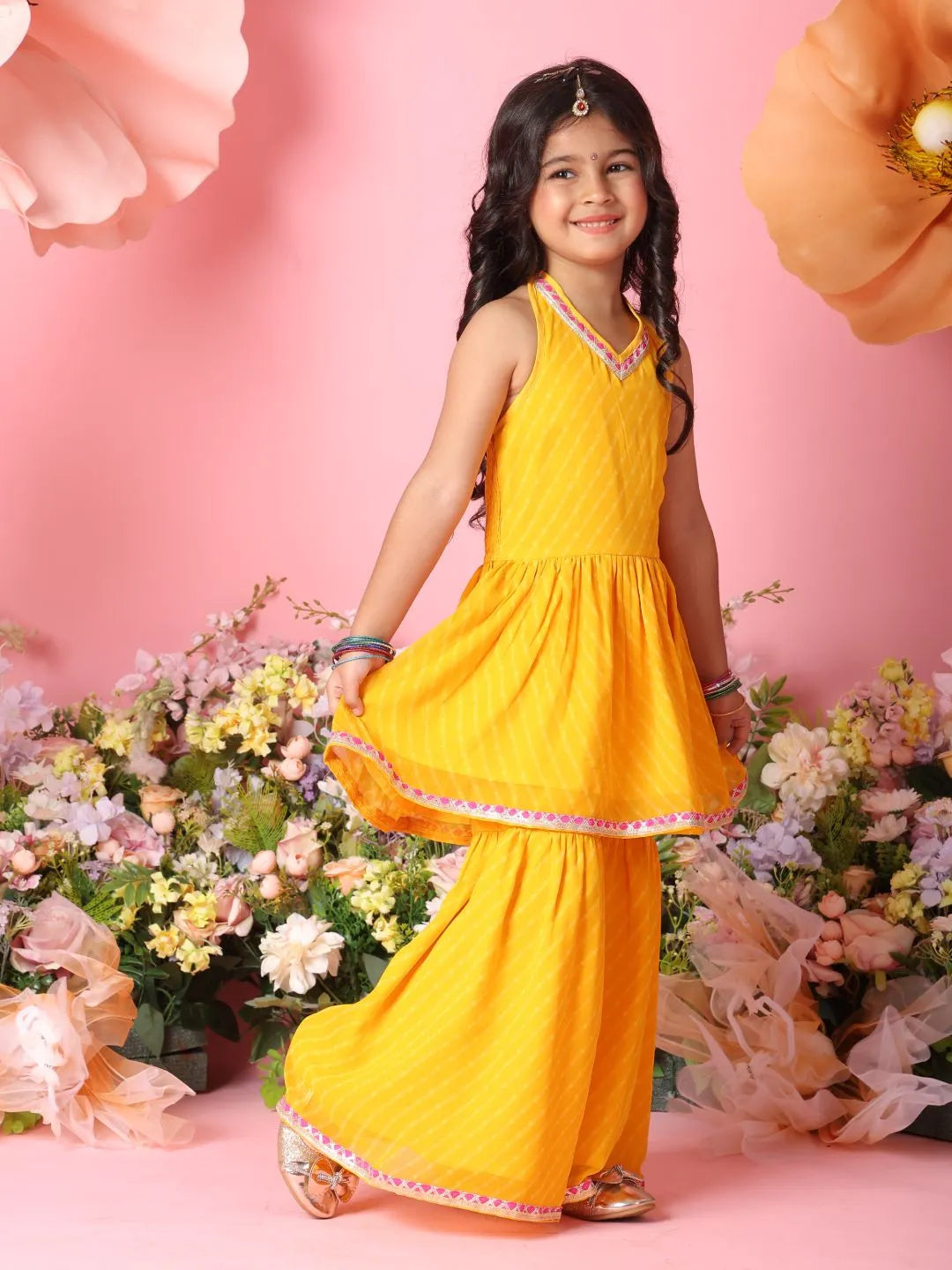 Young girl in a yellow traditional outfit standing among flowers on a pink background