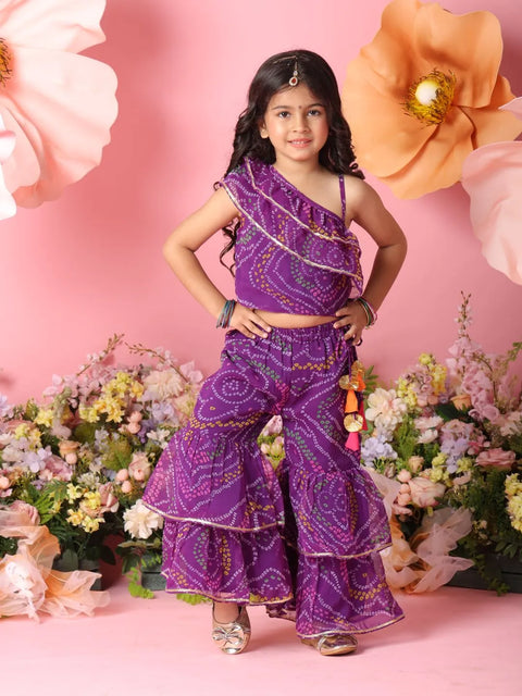Young girl in a purple traditional outfit standing against a pink background with flowers.