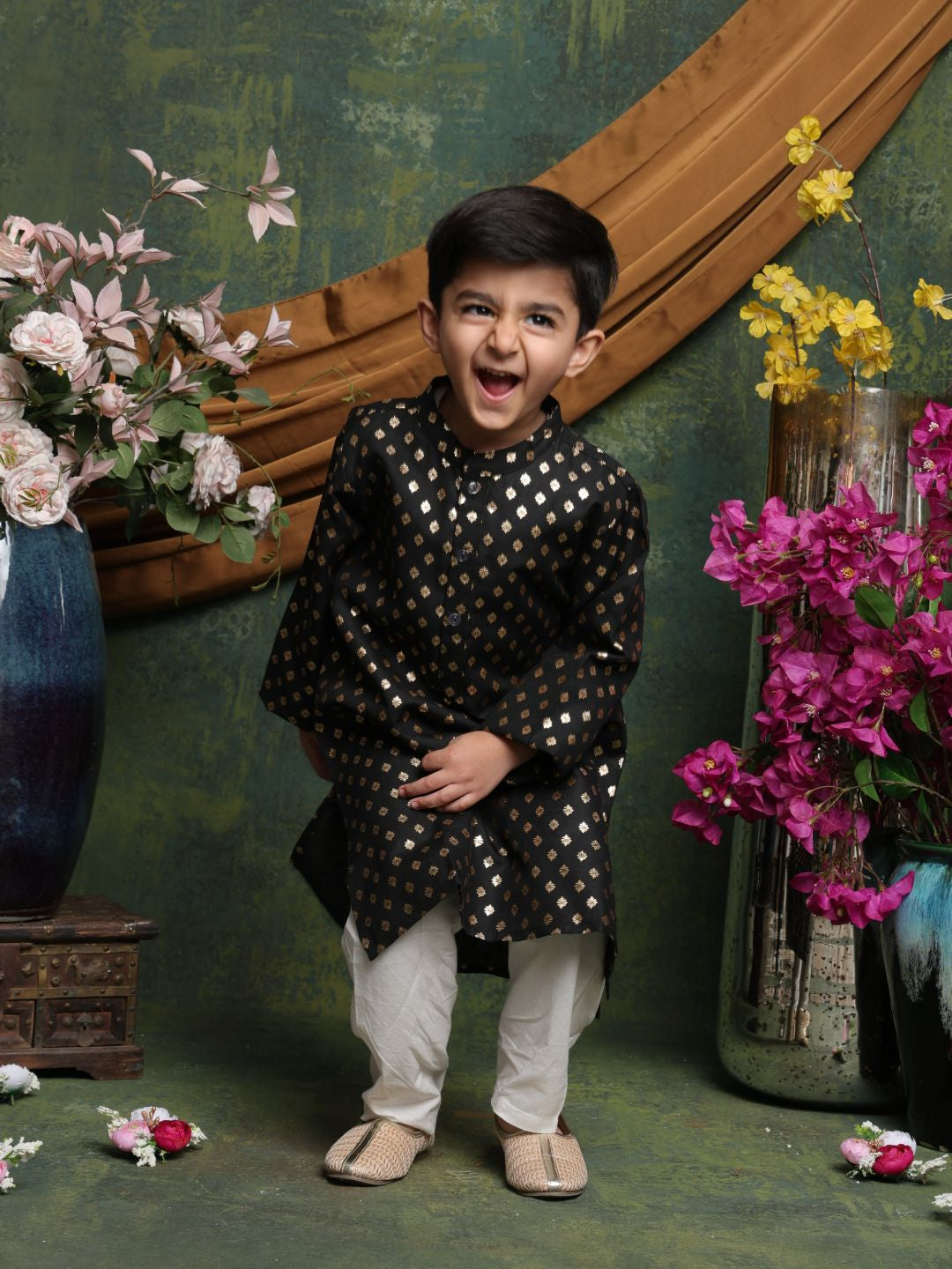 Child in a black and white traditional outfit standing in front of decorative flowers and a wooden wall.