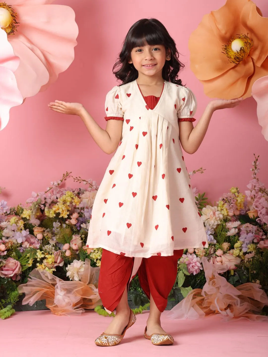 Young girl in a white dress with red heart patterns and red pants holding large flowers against a pink floral background.