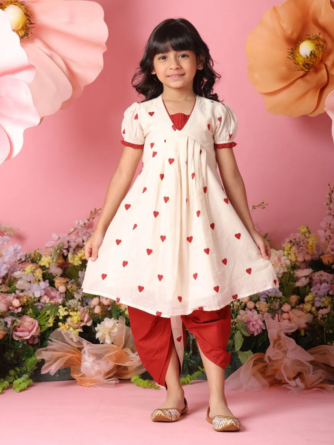Young girl in a white dress with red heart patterns and red pants standing against a pink floral background