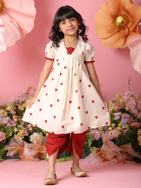 Young girl in a white dress with red hearts and red pants standing against a floral background.