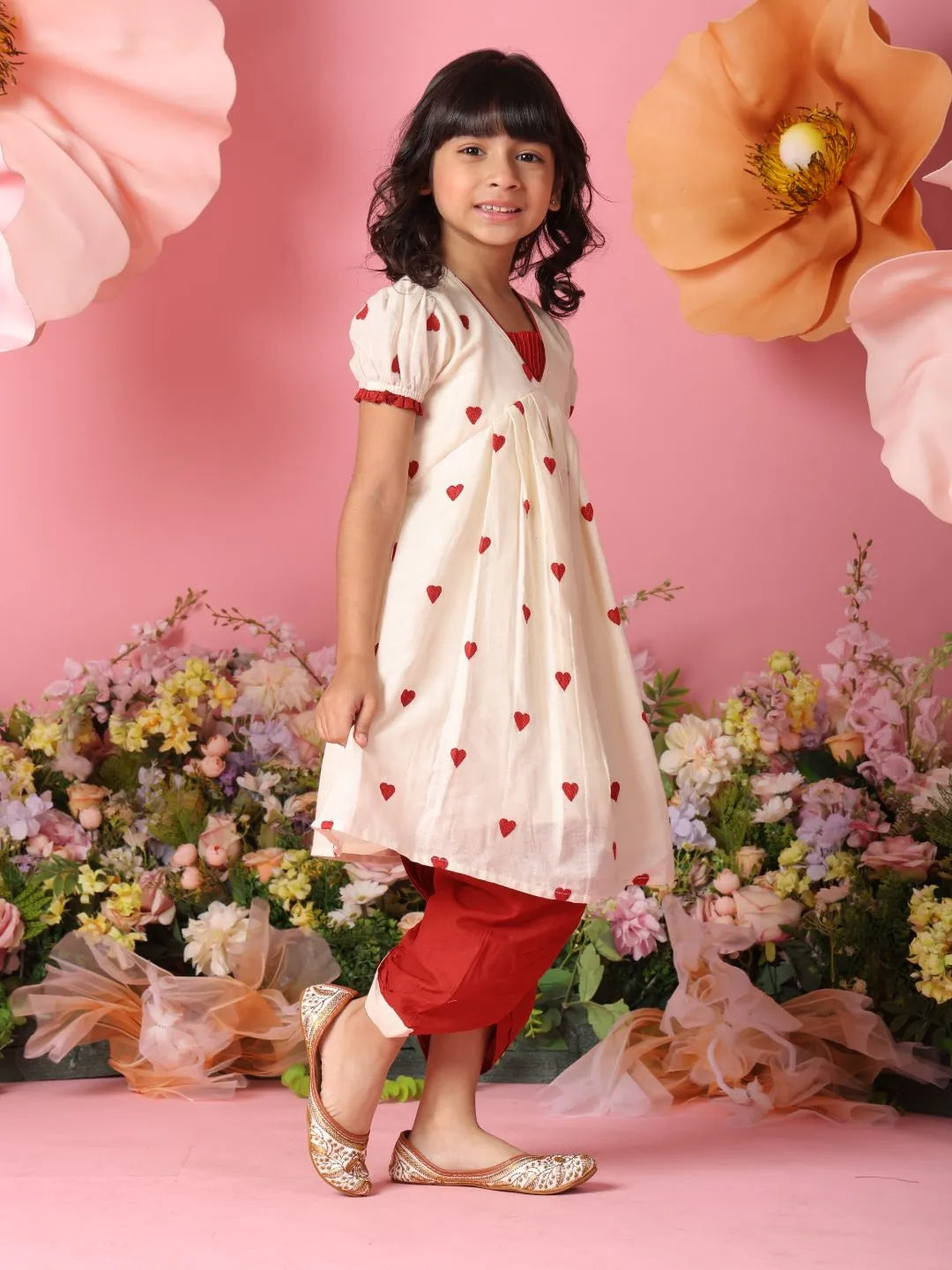 Young girl in a white dress with red heart patterns and red pants standing in front of pink flowers.
