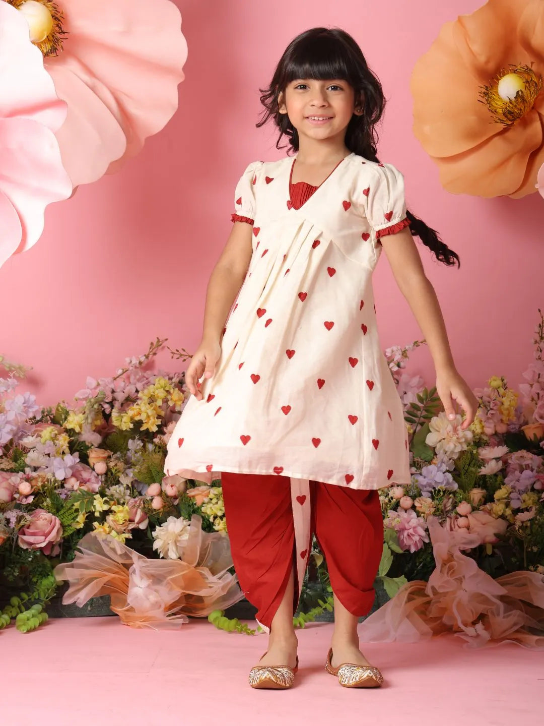 Young girl in a white kurta with red heart patterns and red dhoti standing against a pink floral background
