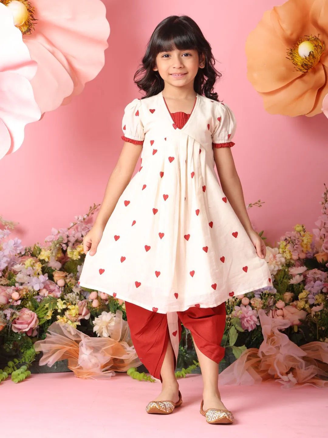 Young girl in a white dress with red heart patterns and red pants standing against a pink floral background.