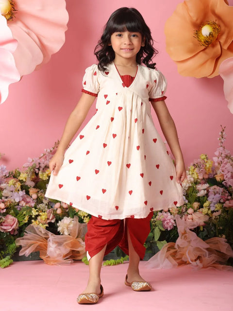 Young girl in a white dress with red heart patterns and red pants standing against a pink floral background.