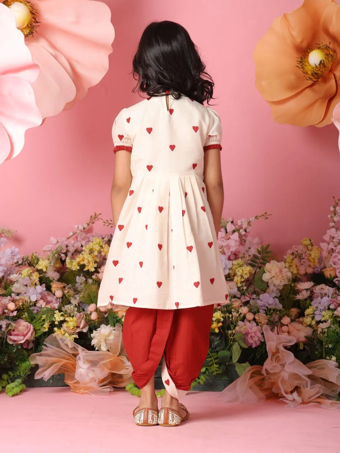 Child wearing a white dress with red heart patterns and red pants against a pink floral background