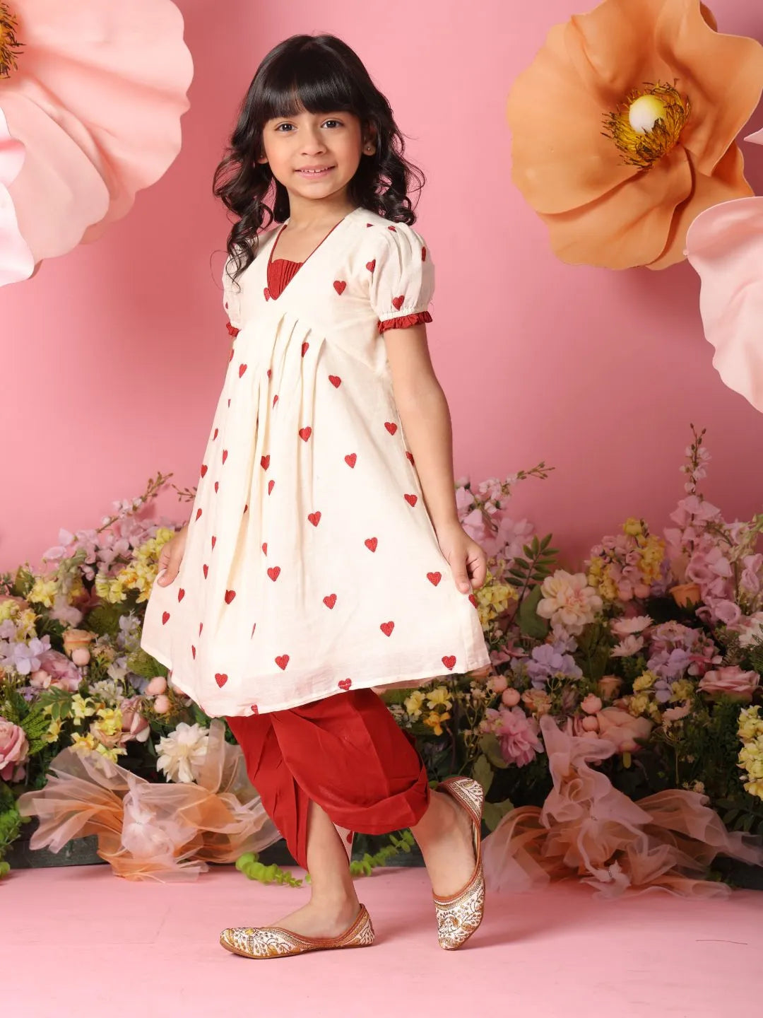 Young girl in a white dress with red hearts and red pants standing in front of flowers and pink background