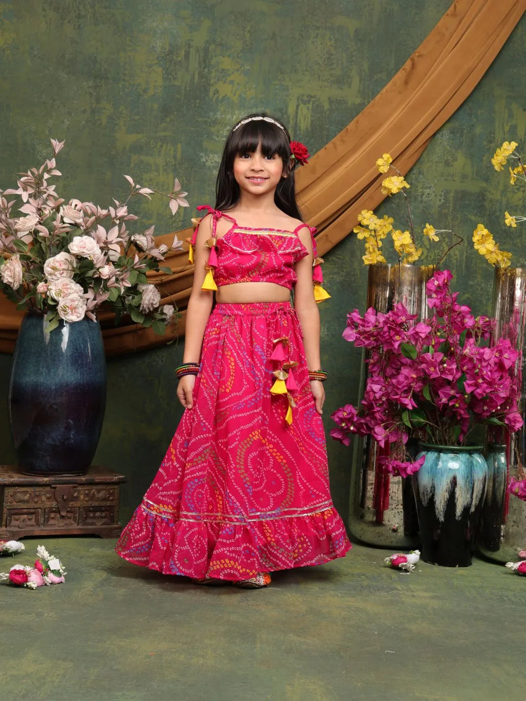 Young girl in a pink traditional outfit standing in front of decorative flowers and vases.