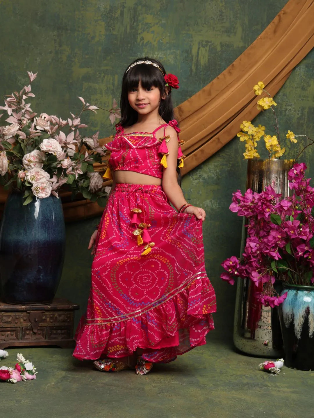 Young girl in a pink traditional outfit standing in front of decorative flowers and a mirror.