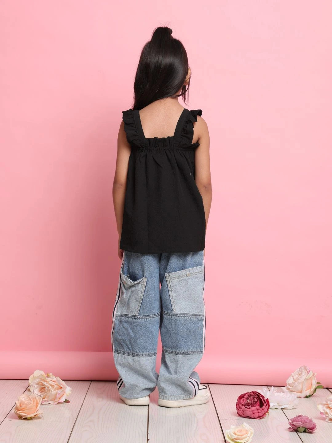 Person wearing a black sleeveless top and blue jeans standing against a pink wall with flowers on the floor.