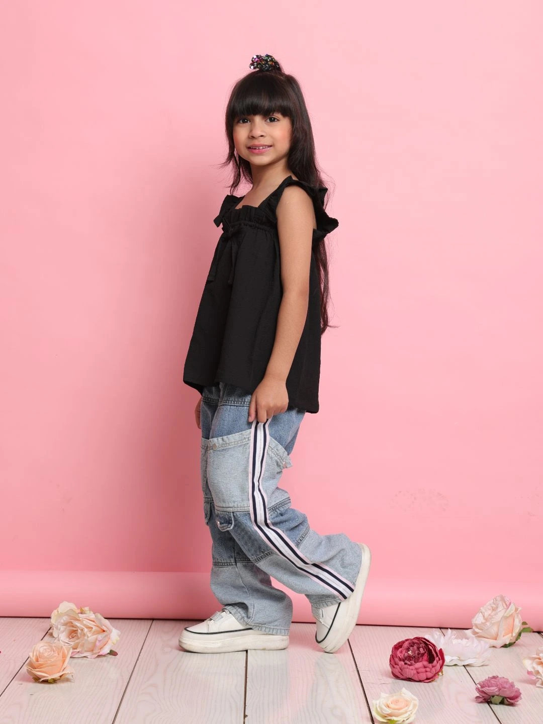 Young girl in black top and blue jeans standing against a pink wall with flowers on the floor.