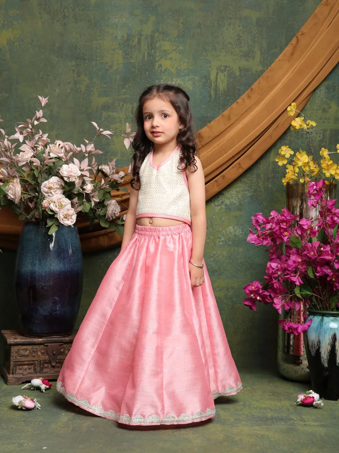 Young girl in a white top and pink skirt standing in front of floral decorations.