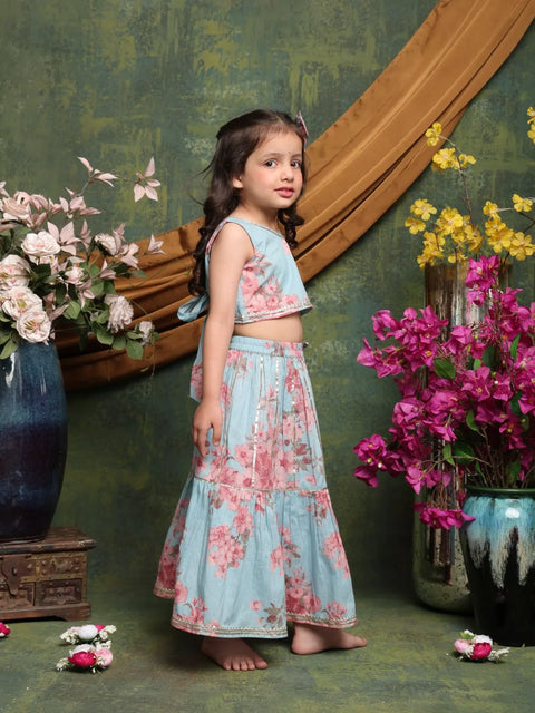 Young girl in a floral dress standing in front of a decorative wall with flowers and vases.