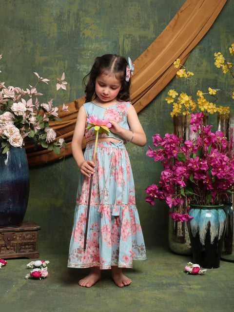 Young girl in a floral dress holding a flower in a decorative setting with flowers and a green wall.