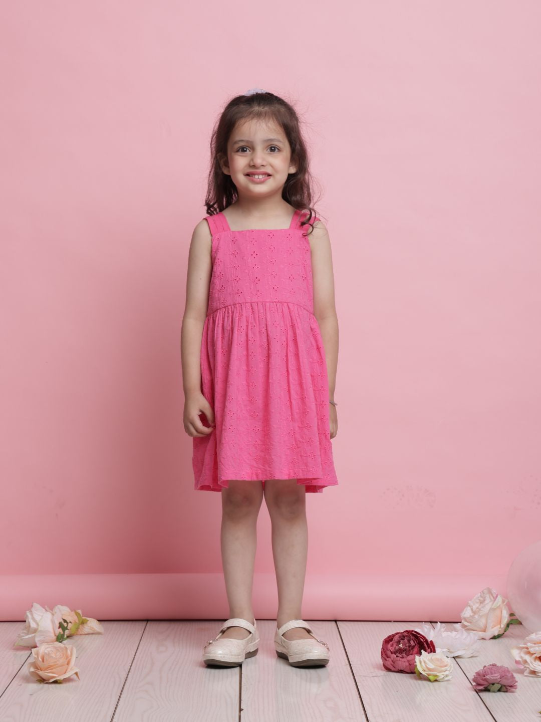 Young girl in a pink dress standing against a pink background with flowers on the floor.