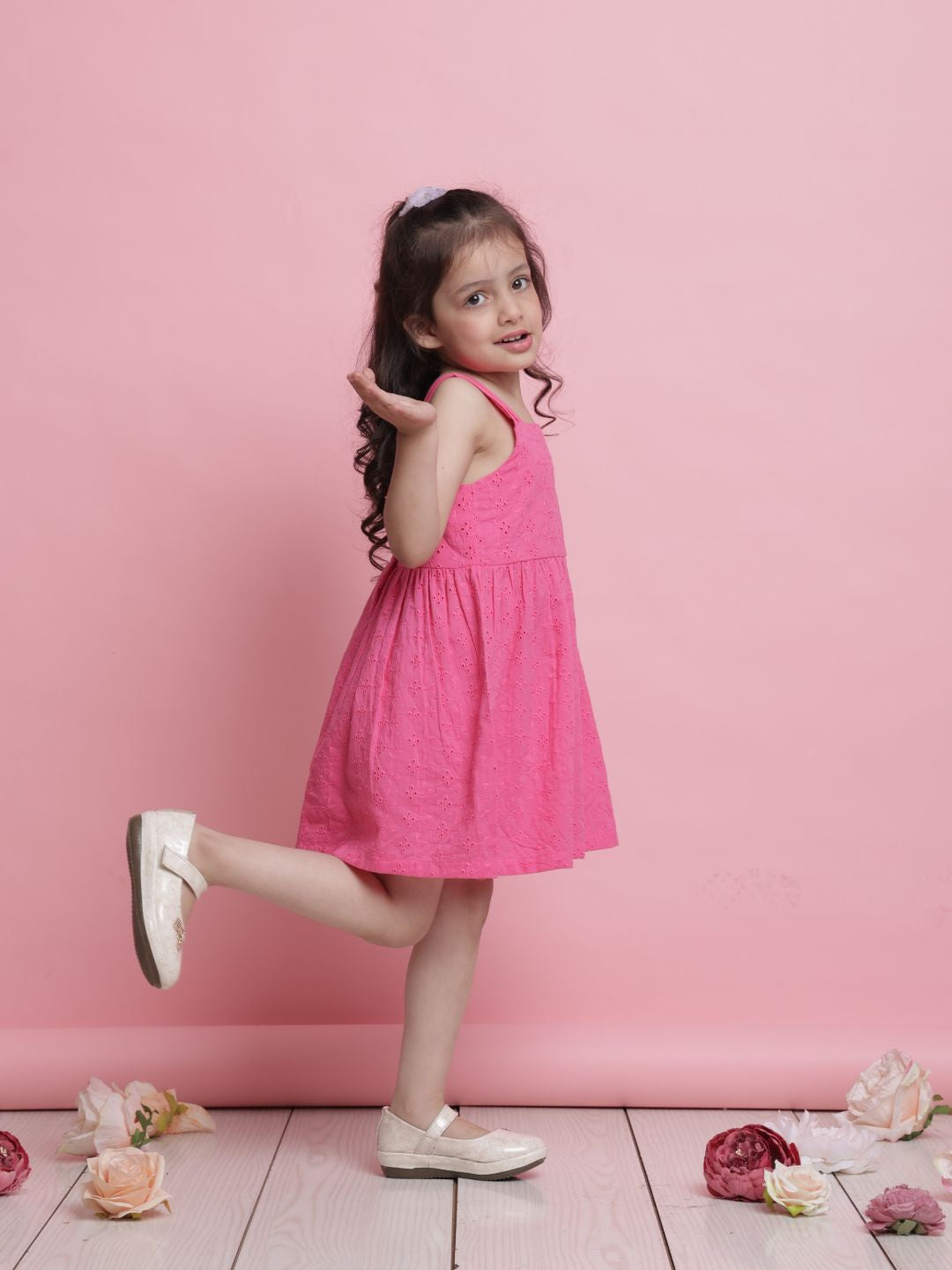 Young girl in a pink dress standing against a pink background with flowers on the floor.