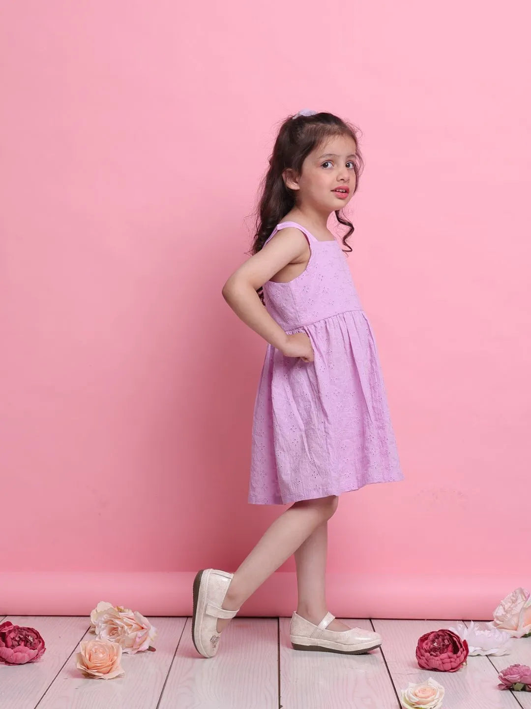 Young girl in a purple dress standing against a pink background with flowers on the floor.