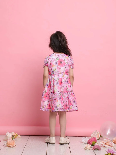 Young girl wearing a floral dress standing against a pink wall with flowers on the floor.