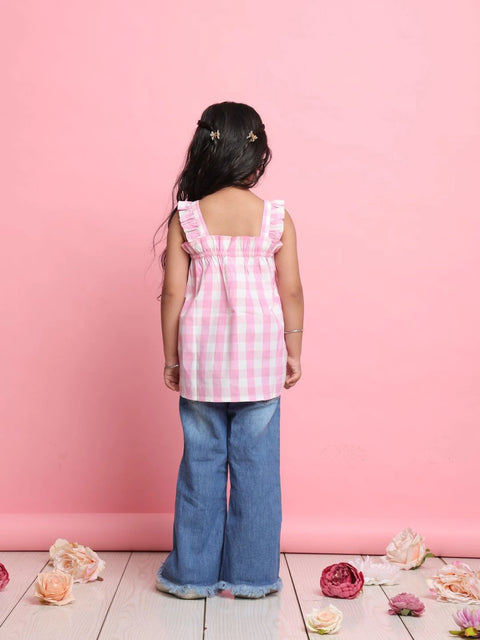Young girl wearing a pink checkered top and blue jeans standing against a pink wall with flowers on the floor.