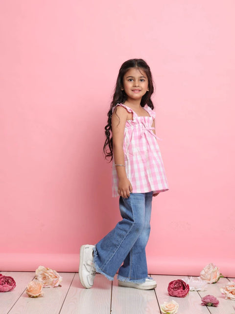 Young girl in a pink checkered top and blue jeans standing against a pink background with flowers on the floor.