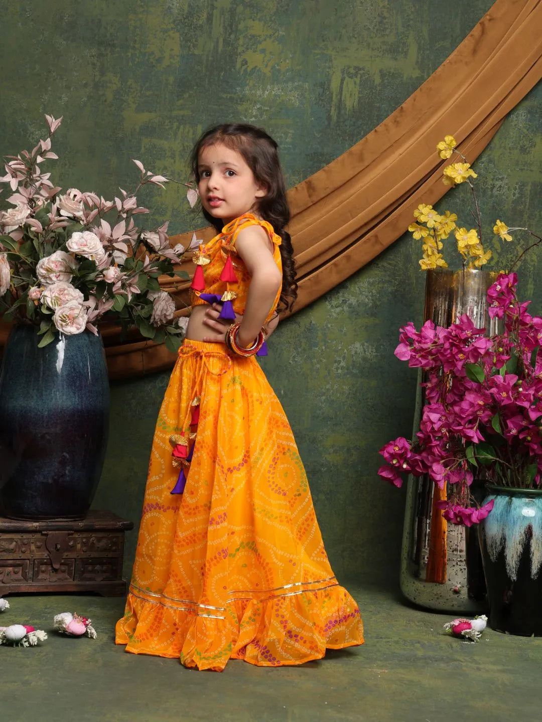 Young girl in a bright orange traditional outfit standing in front of decorative vases with flowers.