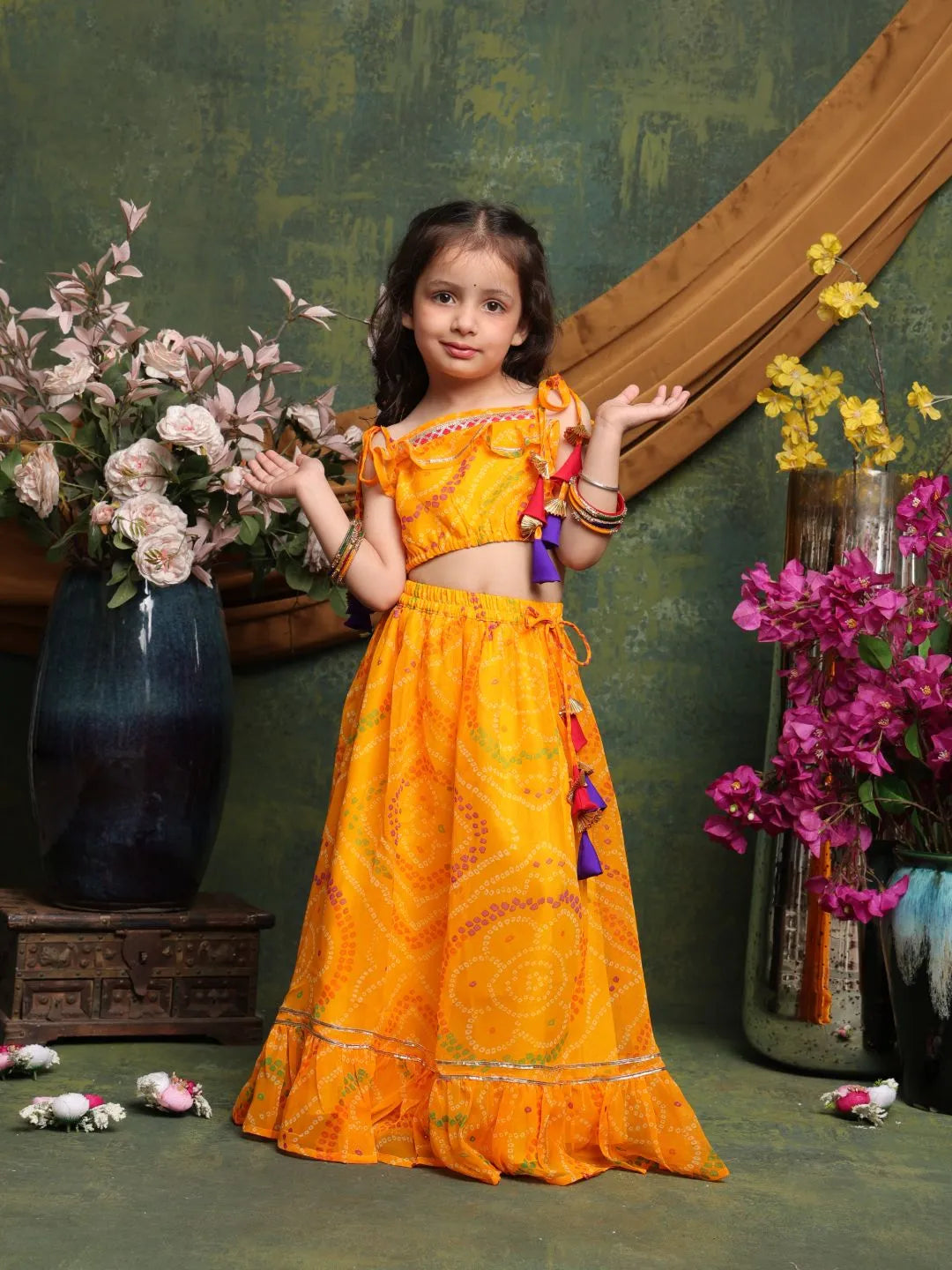 Young girl in a traditional orange outfit standing in front of floral decorations.