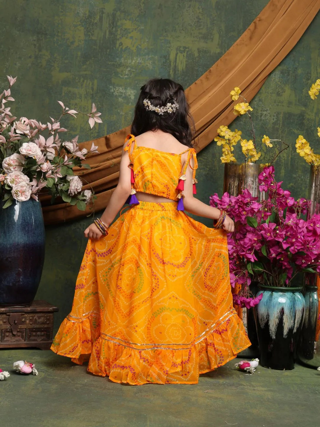Person wearing a bright orange traditional outfit with floral decorations, standing in front of a decorative wall with flowers.
