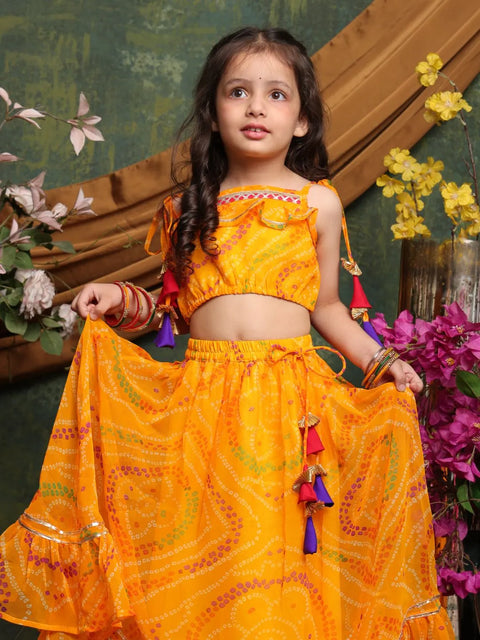 Young girl in a bright orange traditional outfit with colorful tassels, standing against a decorative background with flowers.