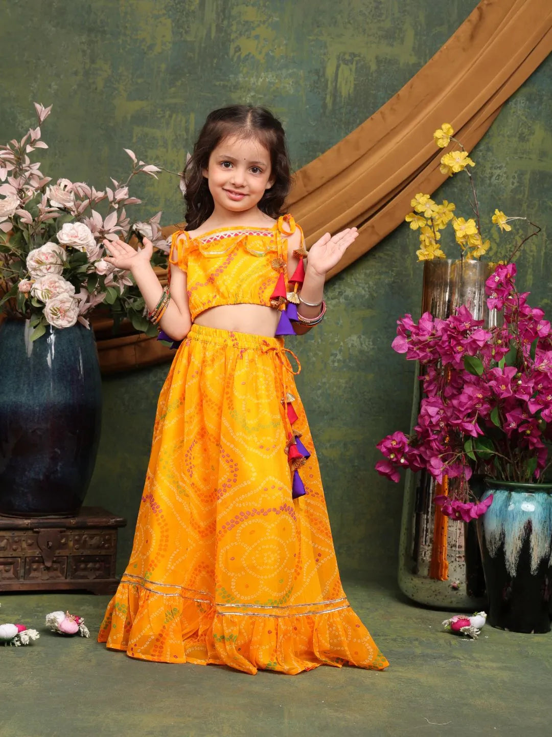 Young girl in a bright orange traditional outfit standing in front of decorative flowers and vases.