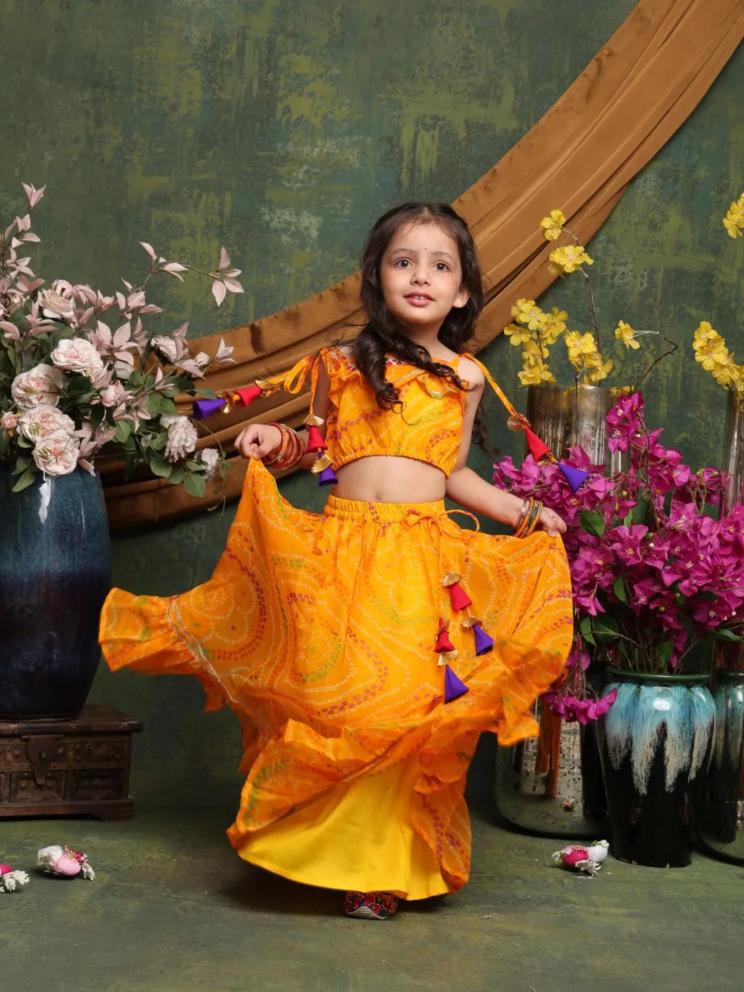 Young girl in a bright yellow traditional outfit with colorful tassels, standing in front of decorative flowers and vases.