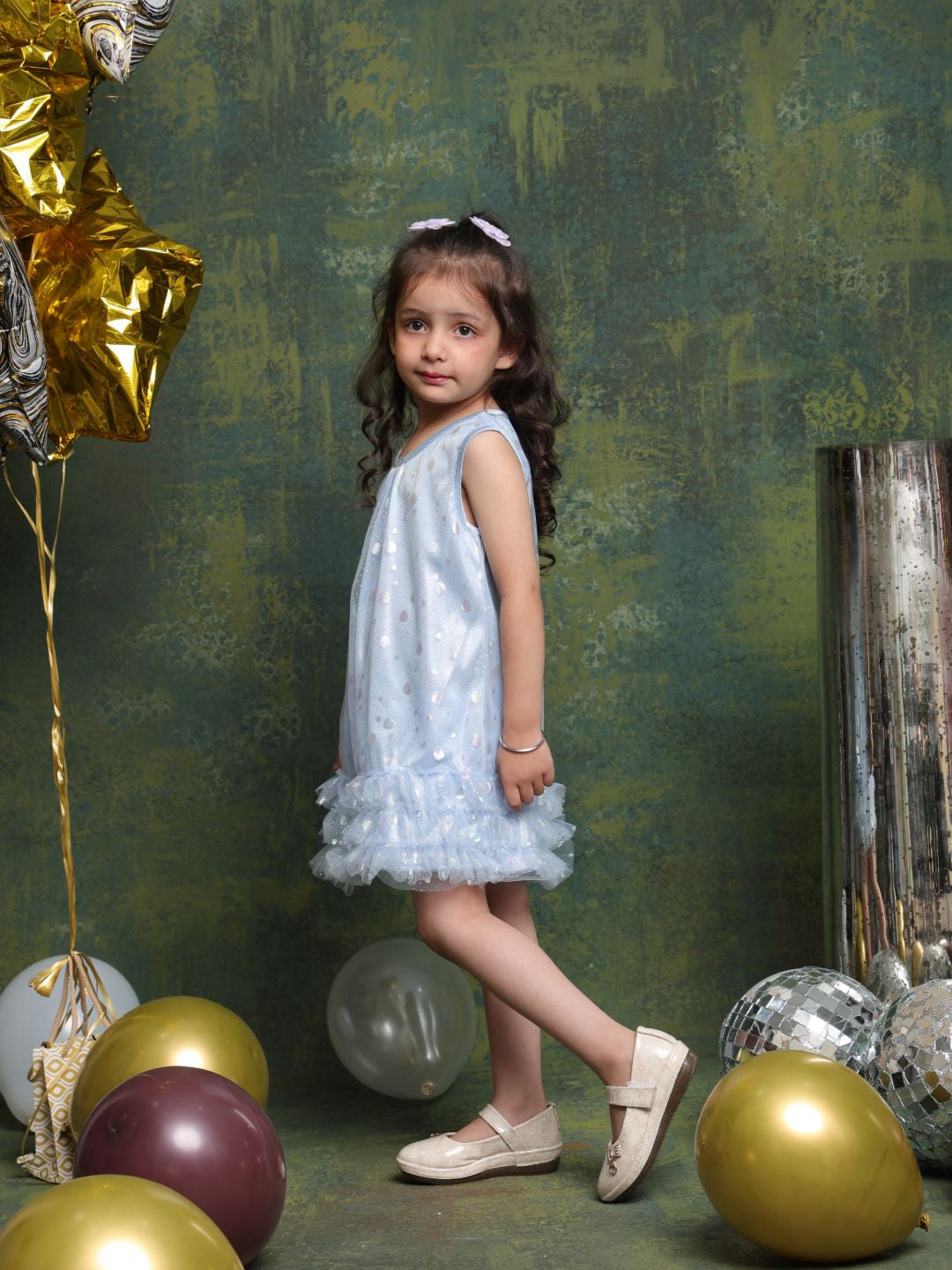Young girl in a light blue dress standing among balloons against a textured green wall.