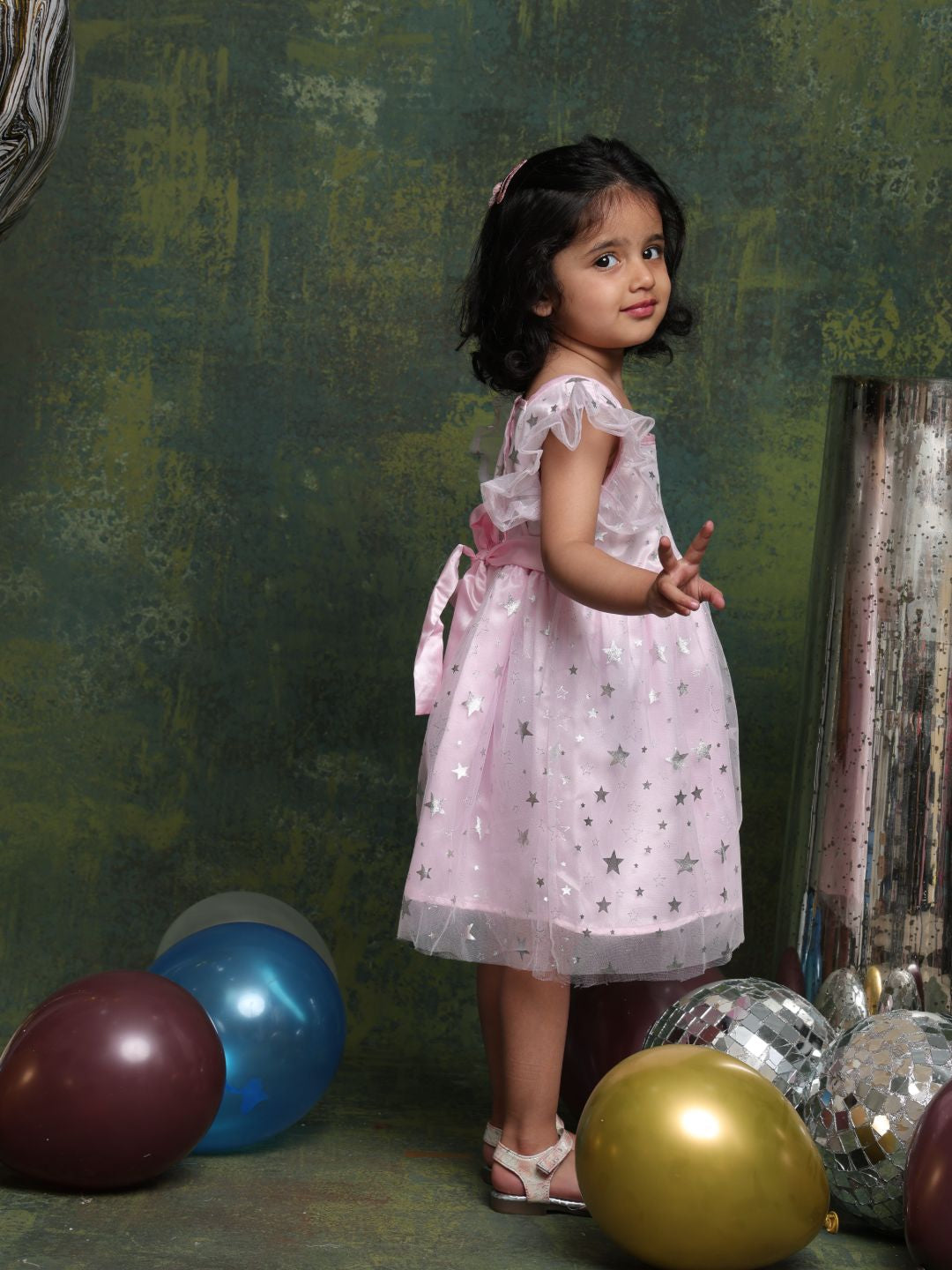 Young girl in a pink dress standing among colorful balloons against a textured green wall.