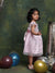 Young girl in a pink dress standing among colorful balloons against a textured green wall.