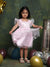 Young girl in a pink dress with white wings standing among colorful balloons against a textured green wall.