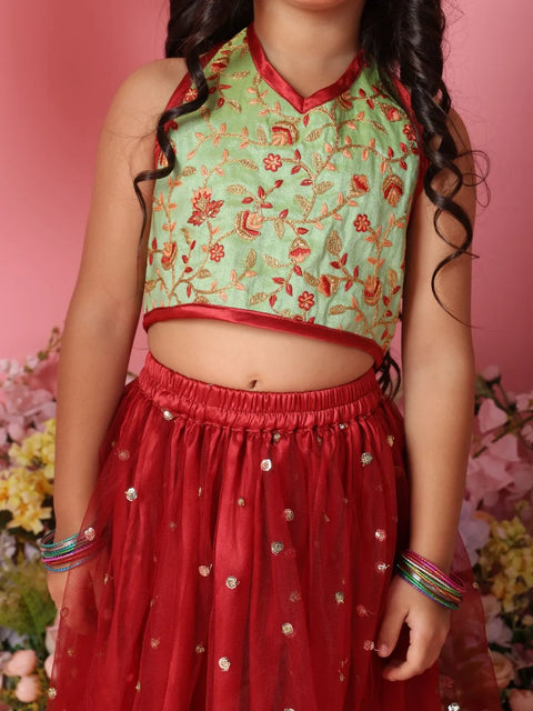 Person wearing a green and red traditional outfit with floral patterns against a pink background.