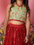Person wearing a green and red traditional outfit with floral patterns against a pink background.