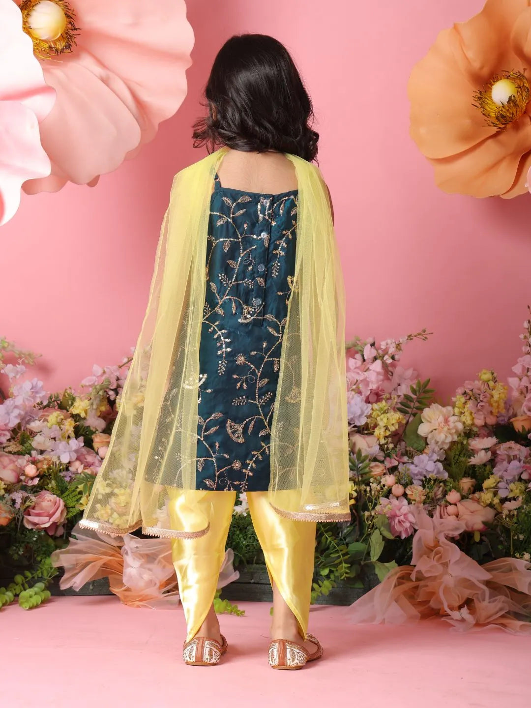 Person wearing a yellow and blue traditional outfit with floral patterns against a pink background with flowers.