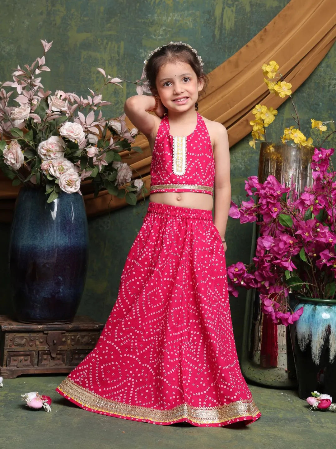 Young girl in a pink traditional outfit standing among flowers and decorative vases.