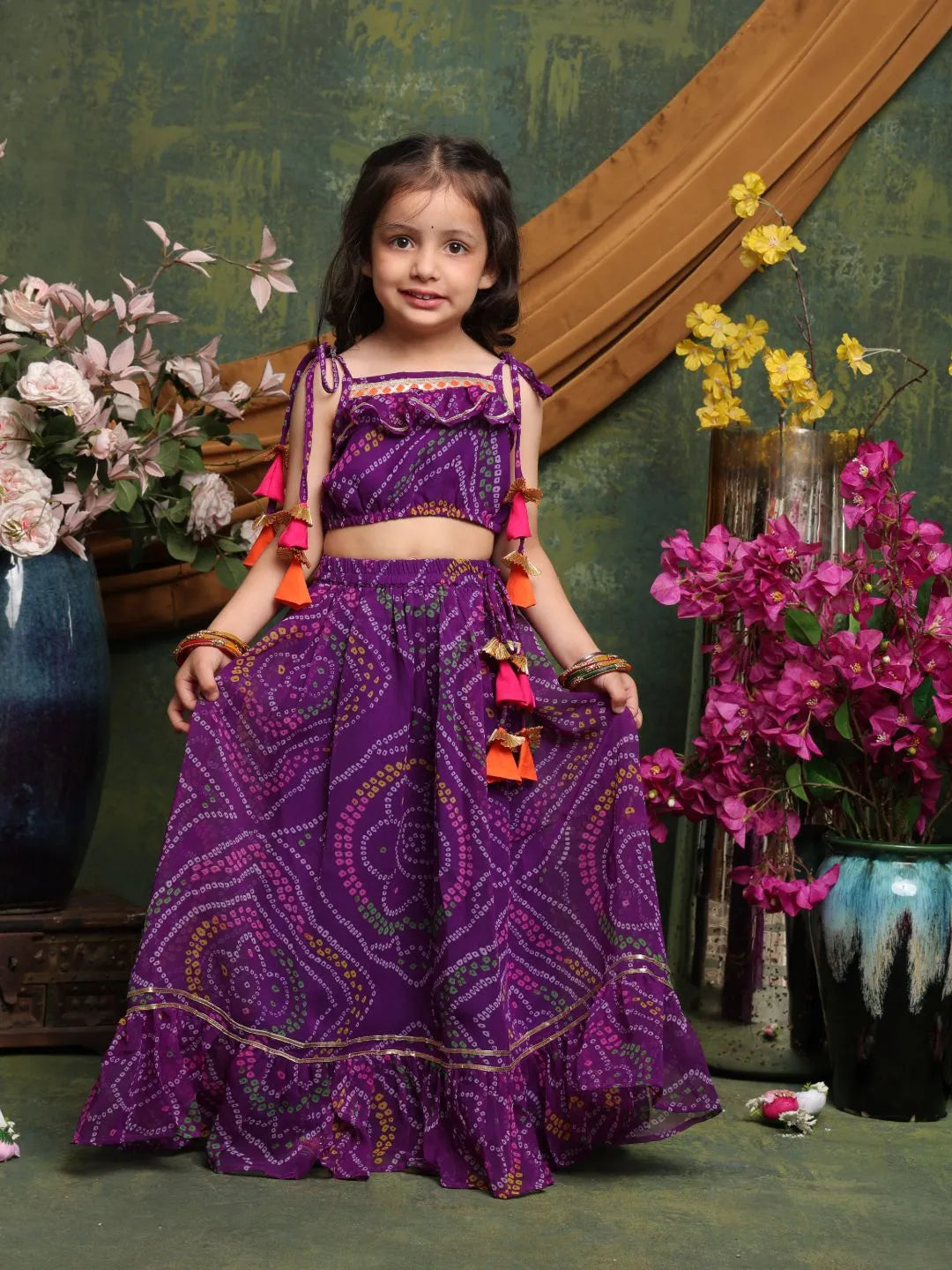 Young girl in a purple traditional outfit with floral decorations, standing against a decorative wall.