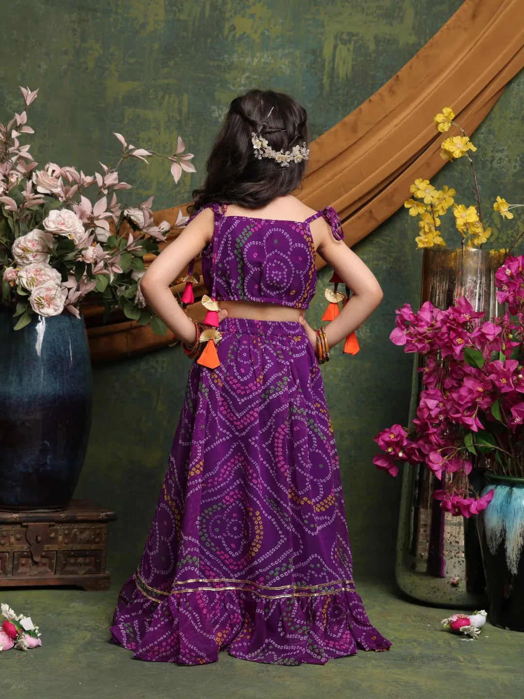 Woman in a purple traditional outfit standing in front of a decorative wall with flowers.