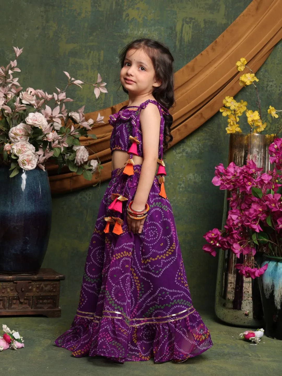 Young girl in a purple traditional outfit standing in front of floral decorations.