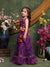 Young girl in a purple traditional outfit standing in front of floral decorations.