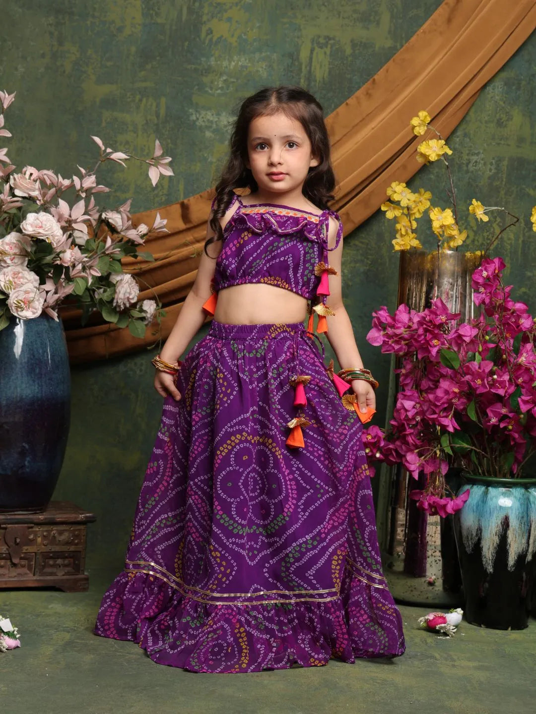 Young girl in a purple traditional outfit standing in front of floral decorations.