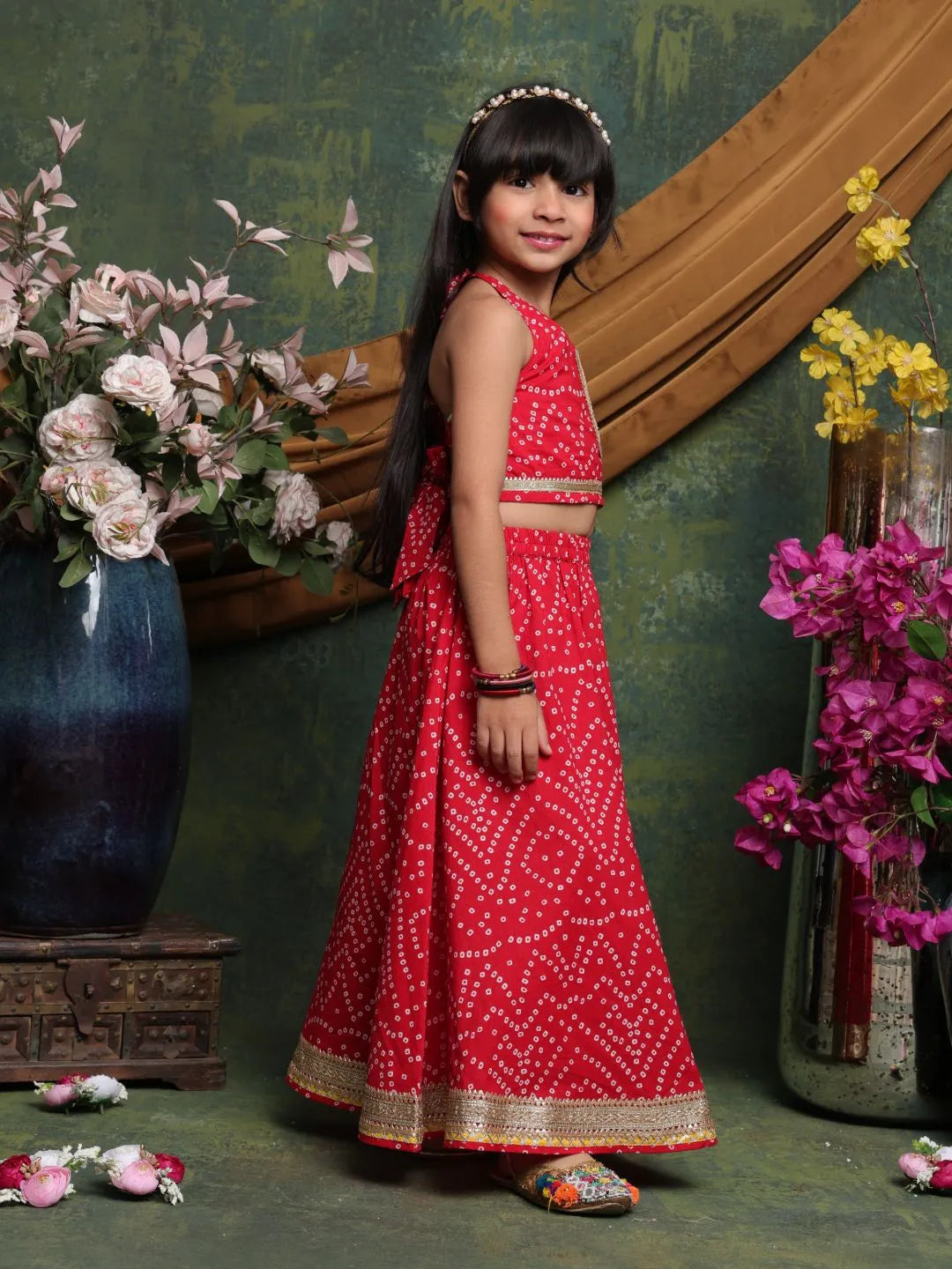 Young girl in a red traditional outfit standing in front of floral decorations.
