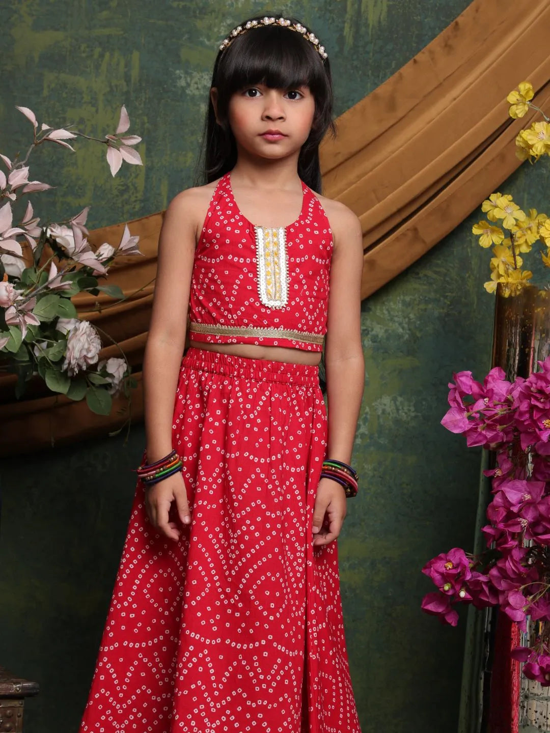 Young girl in a red traditional outfit standing in front of a decorative wall with flowers.