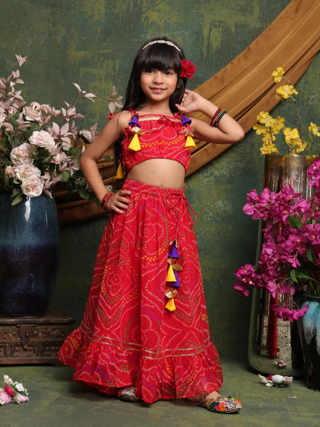 Young girl in a red traditional outfit with colorful decorations, standing against a decorative green wall with flowers.