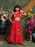 Young girl in a red traditional outfit with colorful decorations, standing against a decorative green wall with flowers.